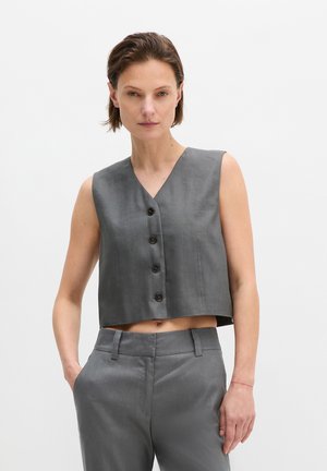 Femme portant un haut court gris sans manches à boutons et un pantalon taille haute assorti, debout avec une main dans la poche devant un fond uni.