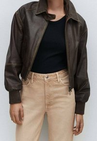 Veste bomber en cuir marron avec fermeture éclair, poignets côtelés et col. Associée à un pantalon beige taille haute et un haut noir côtelé.