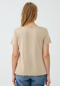 Beżowa koszulka z krótkim rękawem wykonana z gładkiego, lekkiego materiału, z okrągłym dekoltem i prostym wykończeniem, zestawiona z jasnoniebieskimi dżinsami.