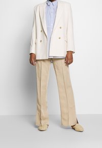 Dubbelknäppt krämfärgad kavaj över en blå och vit randig skjorta, ihop med beigefärgade vida byxor och texturerade beigea slip-on-skor.