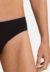 Male torso and upper thigh wearing black briefs with visible stitching and a thin white waistband stripe against a plain background.