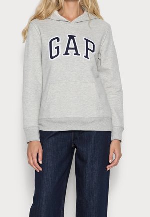 Woman wearing a light gray GAP hoodie with a front pocket and dark blue jeans, standing against a plain background.