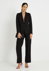 Costume noir sur mesure avec un blazer ajusté et un pantalon à jambes larges. Accessoires comprenant une pochette noire et des sandales à talons hauts roses.