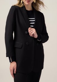 Svart dubbelknäppt kavaj med två runda knappar, skräddarsydd form och fickdetalj. Bärs över en randig svart och vit tröja.