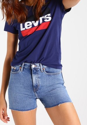 Vrouw draagt een blauwe Levi's T-shirt en een denim short met hoge taille en rafelige zoom, staand tegen een effen achtergrond.