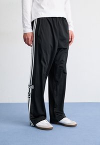 Cilvēks valkā melnas Adidas sporta bikses ar baltām svītrām un baltas kurpes, stāv uz zila grīdas pretī vienkāršai baltai sieni.