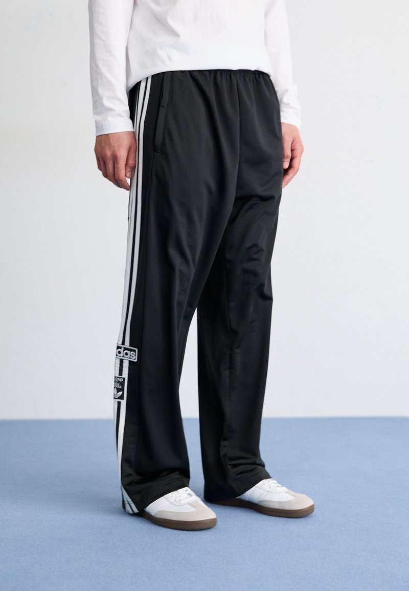 Cilvēks valkā melnas Adidas sporta bikses ar baltām svītrām un baltas kurpes, stāv uz zila grīdas pretī vienkāršai baltai sieni.