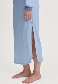 Langes blaues Kleid mit horizontalen Streifen, seitlichem Schlitz und gerippten Bündchen. Der Stoff wirkt weich und leicht. Das Modell zeigt barfüßig.