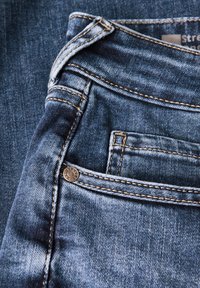 Tmavě modré džíny z denimu s viditelným zapínáním na knoflík, kontrastními švy a malou kapsičkou na mince na pravé straně.