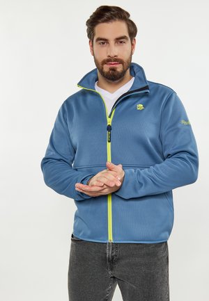 Mann mit Bart, der eine blaue Jacke mit gelbem Reißverschluss und graue Hose trägt, steht mit verschränkten Händen vor einem weißen Hintergrund.