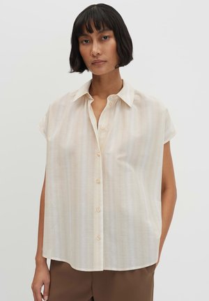 Jonge vrouw met kort zwart haar die een losse, beige blouse met korte mouwen en knopen draagt en een bruine broek tegen een effen achtergrond.