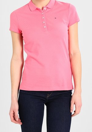 Lyserød polo med krave, korte ærmer og knaplukning. Har en logo på venstre bryst. Kombineret med mørke jeans.