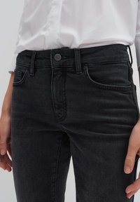 Jean en denim noir avec une coupe slim, présentant une taille à boutons, des poches avant et des détails de couture discrets le long des coutures.
