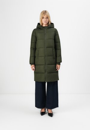 Femme portant un long manteau matelassé à capuche vert olive, un jean sombre à jambes larges et des escarpins noirs à bout pointu, se tenant devant un fond blanc.