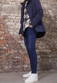 Marinblått isolerat jacka över en rutmönstrad skjorta, i kombination med slim-fit mörka denimjeans och vita plattformssneakers. Tegelvägg i bakgrunden.