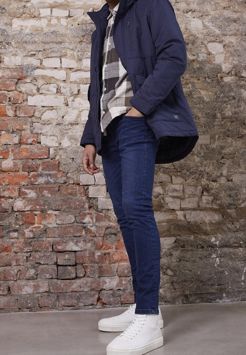 Marinblått isolerat jacka över en rutmönstrad skjorta, i kombination med slim-fit mörka denimjeans och vita plattformssneakers. Tegelvägg i bakgrunden.