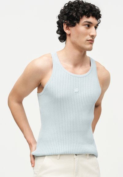 Άνδρας με σγουρά σκούρα μαλλιά, φοράει ένα ανοιχτό μπλε πλεκτό μπλουζάκι χωρίς μανίκια και κρεμ παντελόνι, κοιτάζοντας δεξιά με τα χέρια πίσω από την πλάτη.