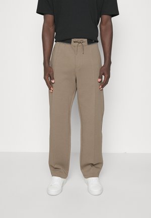 Mann, der beigerweite Hose mit vorderen Nähten, einem Kordelzug in der Taille, einem schwarzen eingesteckten Hemd und weißen Sneakern vor einem einfarbigen Hintergrund trägt.