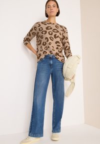 Maglione beige con stampa leopardata, jeans blu a gamba larga e sneakers beige. La modella tiene una borsa color khaki chiaro. Sfondo pulito con elementi minimi.