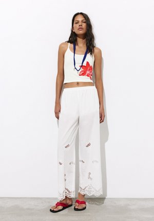 Femme portant un pantalon blanc à jambes larges avec des découpes en dentelle, un crop top blanc avec une fleur rouge, des sandales rouges et un collier de perles bleu.