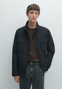 Giacca nera con colletto alto, tasche frontali con bottone a scatto e chiusura a bottone. Indossata sopra un maglione marrone a trama e jeans grigi.