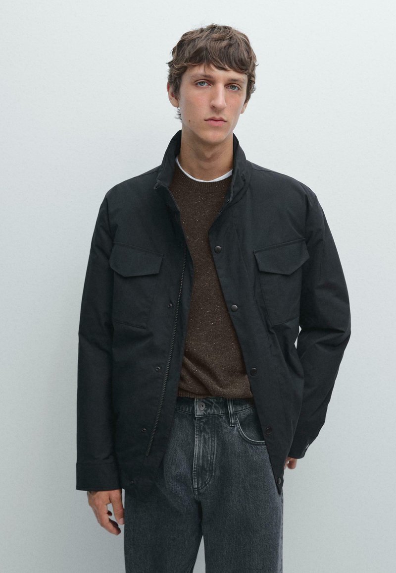 Giacca nera con colletto alto, tasche frontali con bottone a scatto e chiusura a bottone. Indossata sopra un maglione marrone a trama e jeans grigi.