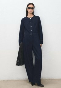 Mono de mezclilla azul marino con mangas largas, cuello redondo, cierre frontal de botones y piernas anchas. Estilizado con un bolso negro con flecos y gafas de sol.