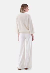 Eine Frau, die seitlich blickt und einen lockeren weißen Strickpullover mit Dreiviertelärmeln sowie weiten weißen Hosen trägt.