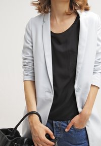 Jasnoszary żakiet z pojedynczym guzikiem, noszony na czarnej koszuli z kieszonką, w zestawie z niebieskimi dżinsami. Czarna torebka z textured detalem.
