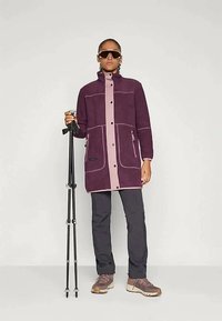 Veste en polaire violette avec un plastron avant rose clair, deux poches latérales, associée à un pantalon sombre et des chaussures de randonnée, tenant des bâtons de trekking.