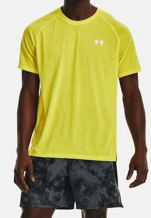 Mężczyzna ubrany w jasnożółtą sportową koszulkę z krótkim rękawem i czarno-szare sportowe szorty w wzory, stojący z rękami swobodnie opuszczonymi wzdłuż ciała.