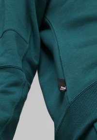Sudadera azul verdosa de tejido suave con puños de canalé. Presenta una etiqueta de logo cerca del dobladillo y detalles de costura en las mangas.