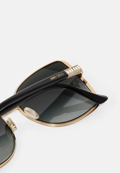 Černé sluneční brýle se zlatým kovovým rámem, zdobené texturovaným akcentem na spánkách. Skla jsou tmavě šedá a poskytují UV ochranu.