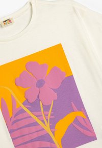 Kerman t-paita, jossa on suuri kukkakuva pinkin ja violetin sävyissä oranssia ja violettia taustaa vasten. Pehmeä puuvillamateriaali, pyöreä kaula-aukko.