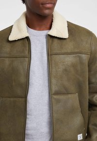 Giacca in similpelle verde oliva con colletto in shearling color crema, chiusura con zip, due tasche frontali e superficie liscia e strutturata.