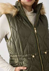 Olive green quilted vest with a faux fur hood, gold zip closure, and drawstring details at the waist. Smooth texture, sleeveless design.