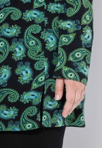 Maglietta nera con motivo paisley verde e blu. Presenta uno spacco laterale e maniche lunghe con risvolto. Tessuto morbido con moderata elasticità.