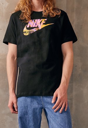 Personne portant un t-shirt noir Nike avec un logo coloré et un jean bleu clair, debout contre un mur en béton uni.
