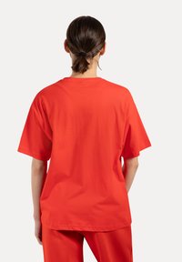 Persona con i capelli castani raccolti, che indossa una maglietta rossa a maniche corte larga e un paio di pantaloni rossi abbinati, che sta in piedi con la schiena visibile.