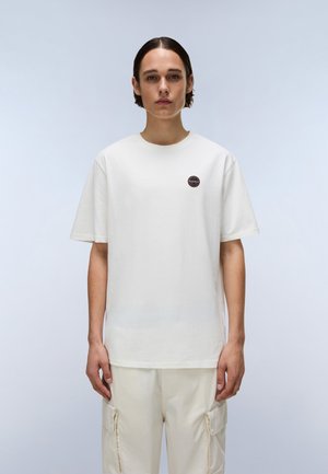 Junge Person mit kurzen dunklen Haaren, die ein schlichtes weißes T-Shirt mit einem kleinen runden Aufnäher auf der linken Brust und hellfarbene Cargohosen trägt.