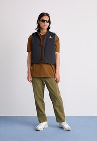 Black sleeveless vest with two zippered pockets and shoulder accents, worn over a brown t-shirt, paired with olive green pants and white sneakers.