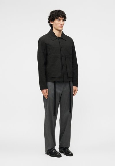 Göndör hajú férfi áll, texturált fekete patentos kabátot, szürke pliszírozott nadrágot fekete pántokkal és fekete bőrcipőt visel.