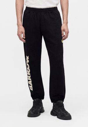 Personne portant un pantalon de survêtement noir avec le texte BARROW sur la jambe gauche et un petit visage souriant sur la cuisse droite, associé à des baskets noires.