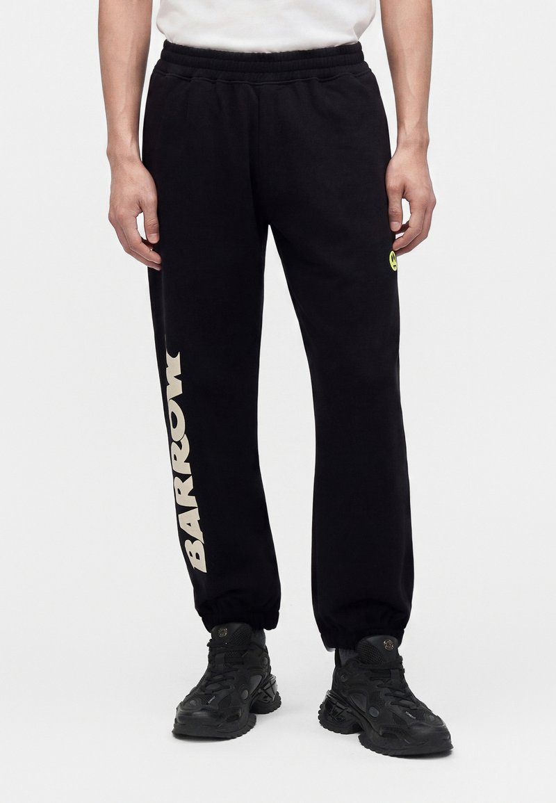Personne portant un pantalon de survêtement noir avec le texte BARROW sur la jambe gauche et un petit visage souriant sur la cuisse droite, associé à des baskets noires.
