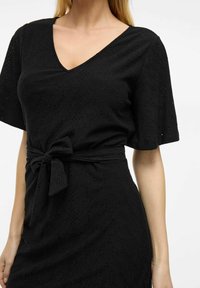 Svart klänning med djup V-ringning, korta ärmar och ett knytbälte i midjan. Har en texturerad tyg med utskärningar och en något lös passform.