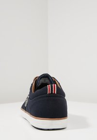 Mörkblå sneaker med vit sula, brun kant och röd-vitrandig dragögla bak, sedd från hälen på en vit yta.