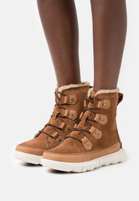 Bottines en suède marron avec doublure en fourrure beige, bout rond et design à lacets avec des accents métalliques et une semelle en caoutchouc blanc robuste.