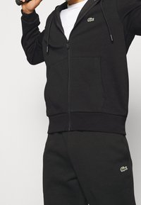 Homme portant un sweat à capuche noir zippé et un pantalon de jogging noir avec des logos de crocodile verts, tenant un haltère noir sur l'épaule.