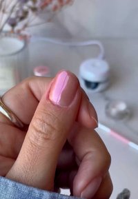 Jasnoróżowy żelowy lakier do paznokci nałożony na kciuk, ukazujący gładkie, błyszczące wykończenie. W tle widoczne rozmyte produkty do paznokci oraz świeca.
