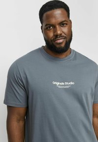 Grå bomulds t-shirt med rund hals, der har et hvidt logo "Originals Studio" trykt på forsiden. Korte ærmer og glat tekstur.